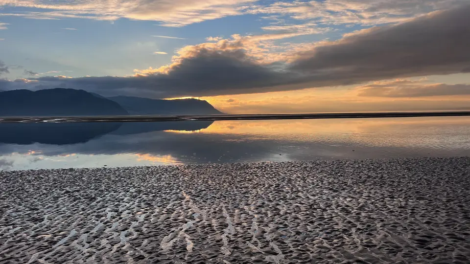 Rippled sand and still water at low tide, reflecting a sunset behind the hills