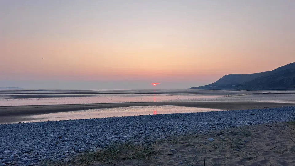 The sun setting over the sea at low tide, with pebbles and dunes in front and a headland on the right