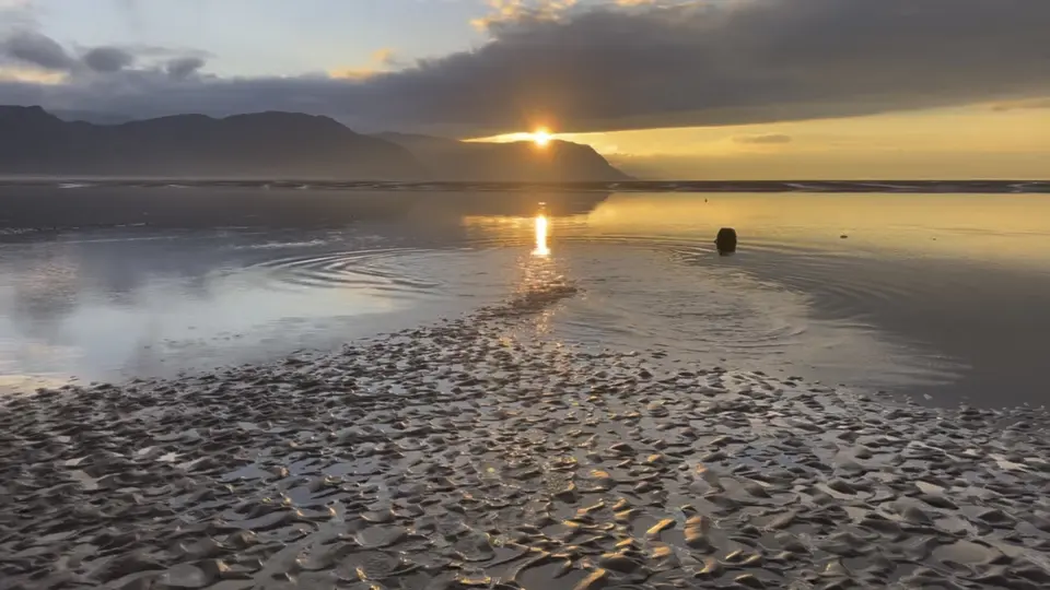 The sun setting behind the hills, reflected in a pool on the rippled sand