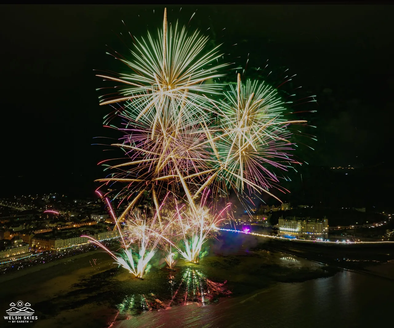 Aerial photograph of the Llandudno fireworks bursting in gold, green and pink over North Shore Beach at night, with the lit-up promenade, the pier and the Grand Hotel behind
